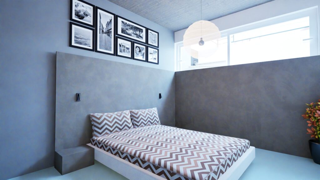 A modern bedroom interior features a low platform bed with a zigzag patterned duvet and pillows, set against a textured gray wall. Above the bed, a collection of framed black and white photographs adorns a blue wall. A simple white pendant light hangs from the ceiling, illuminating the space near a window with a view of a building exterior. The room has a minimalist aesthetic with concrete bedside tables and a potted plant adding a touch of color. By Modify Architects